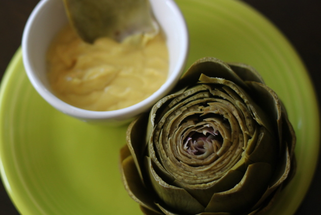 A cooked artichoke on a plate next to a bowl of dipping sauce