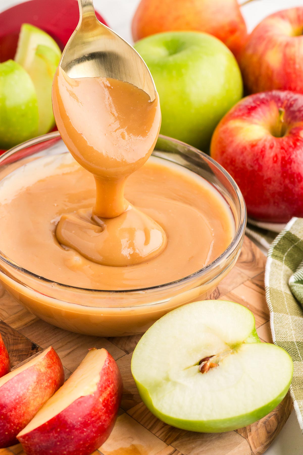 a spoon pouring caramel apple dip into a glass bowl with apples in the background