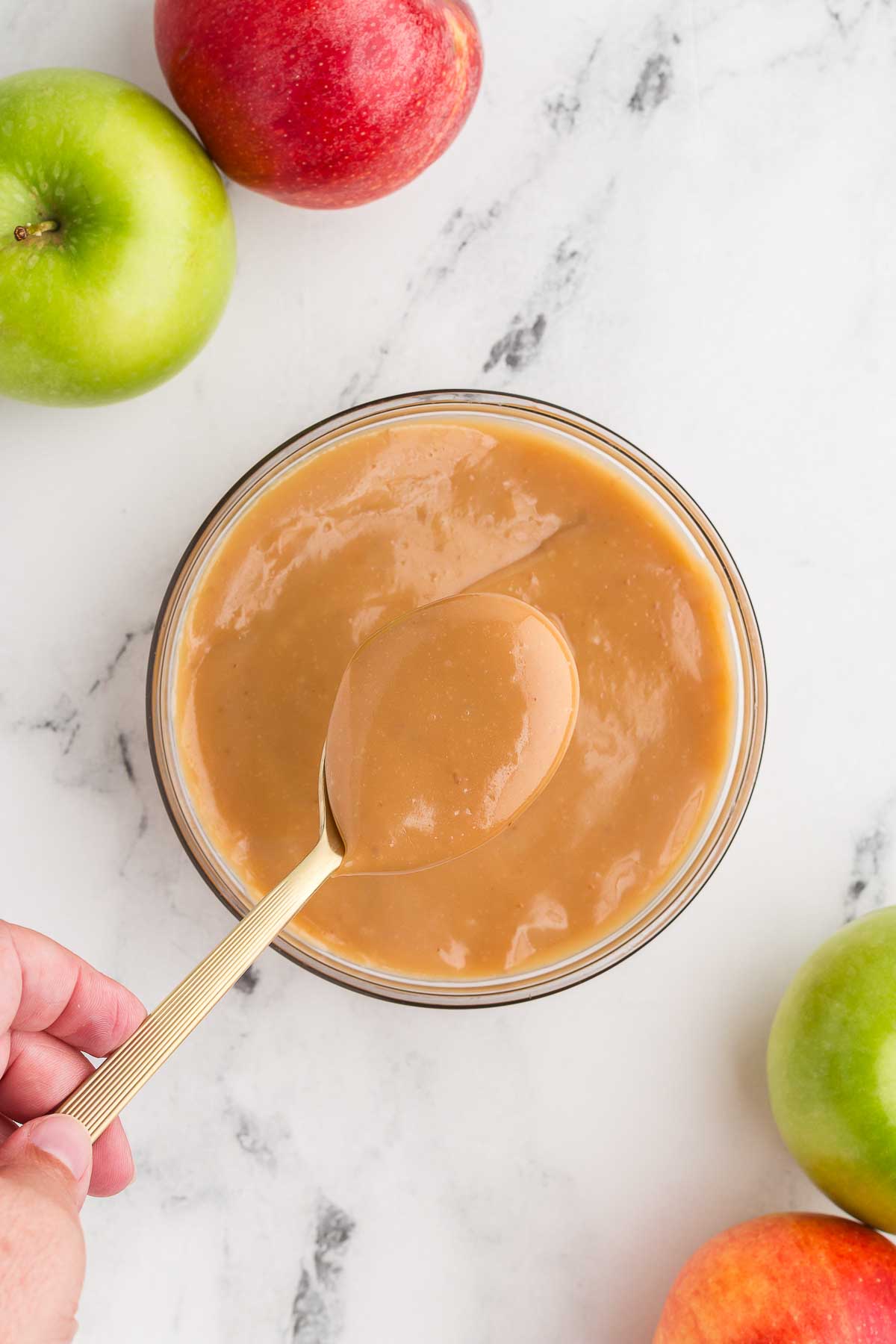 Top view of caramel apple dip in a glass bowl with a spoon lifting up some of the dip and apples in the background