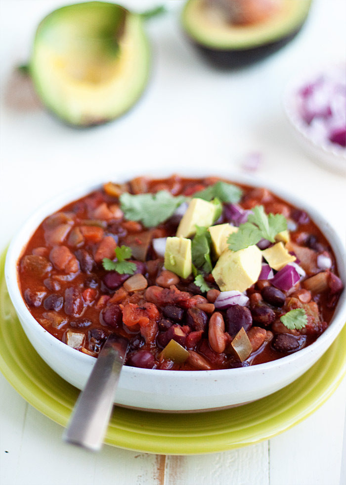 A bowl of 3-bean chili in a white bowl on a lime green plate with avocados, red onion, and cilantro on top. 