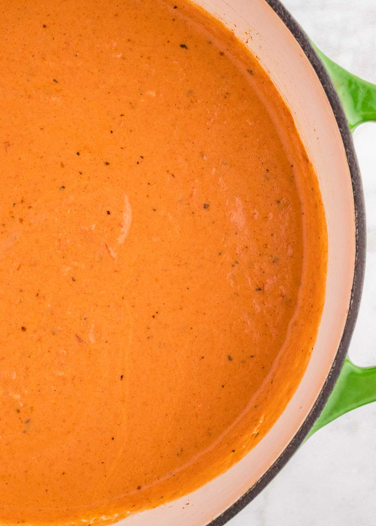 A close-up of creamy tomato soup in a pot with green handles on a marble background.