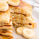A wedge is cut out of fluffy banana pancakes to show how fluffy they are. Slices of bananas and a drizzle of pure maple syrup goes down the side. Banana slices decorate the pancakes and plate.