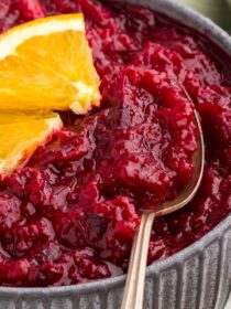 orange cranberry sauce in a gray bowl with a silver spoon and orange garnish
