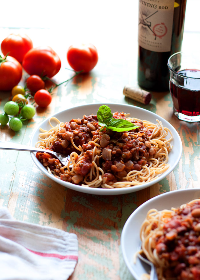 A white pasta bowl full of vegetarian bolognese on top of whole wheat spaghetti. In the background are tomatoes on the vine and a glass of red wine. 