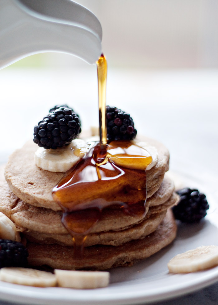 A pile of vegan banana oatmeal blender pancakes with syrup being poured over the top. They are garnished with blackberries and sliced bananas. 