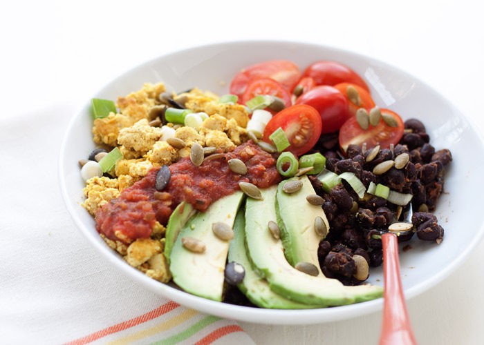 A high protein vegan breakfast bowl in a white bowl with a white background and colorful striped napkin. The bowl contains black beans, tofu scramble, avocados, cherry tomatoes, salsa, and pepitas. 