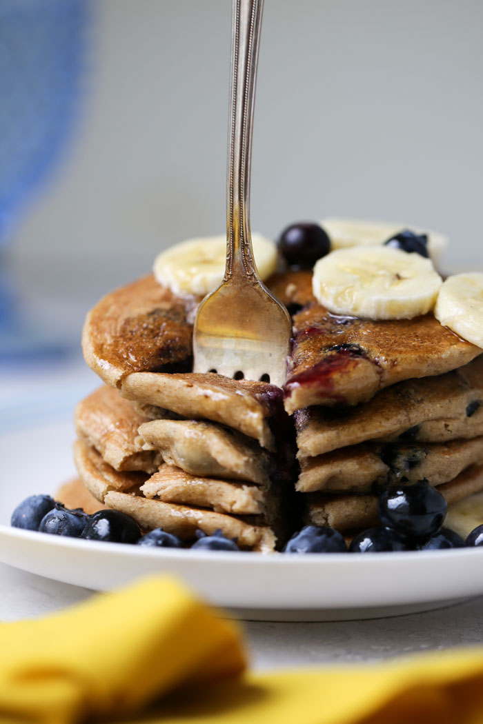 A pile of banana oatmeal pancakes with blueberries and sliced banana. They are drizzled with syrup and have a bite cut out and ready to eat, with a fork stuck in them. 