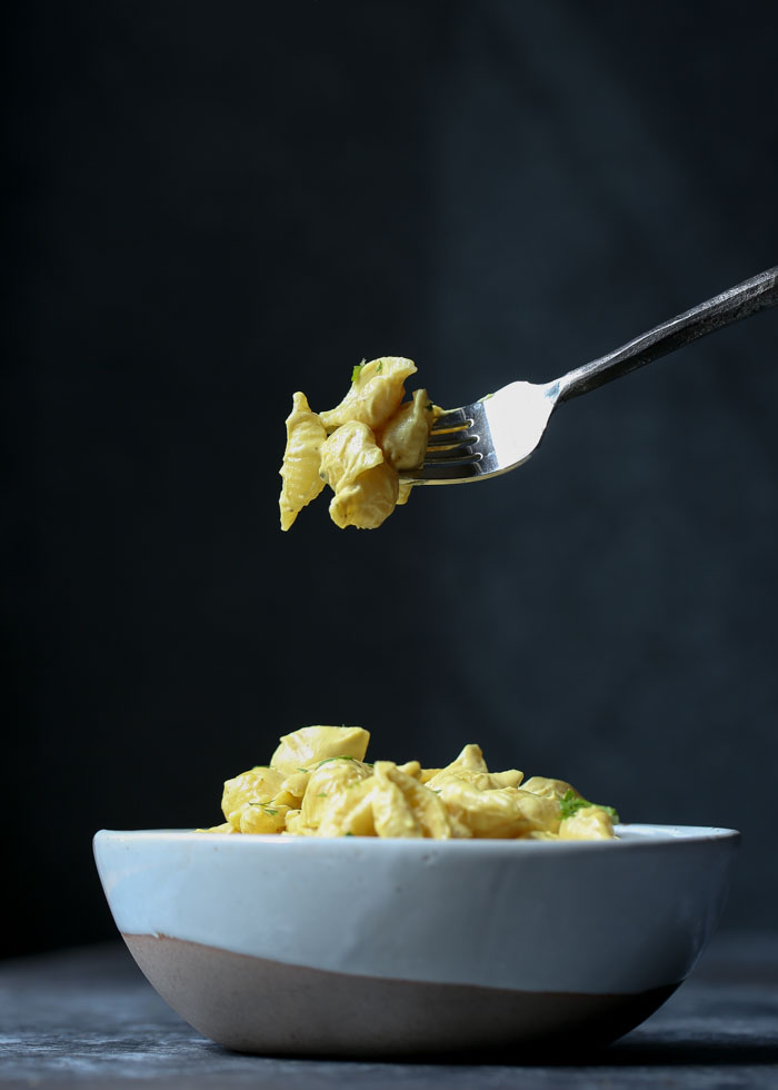 A forkful of dairy free Mac and cheese above a white bowl with more Mac and cheese in front of a black background. 