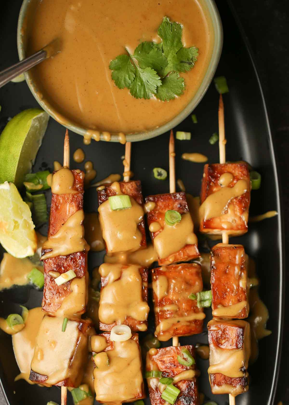 A bowl of peanut sauce above some tofu satay skewers drizzled with more peanut sauce. 