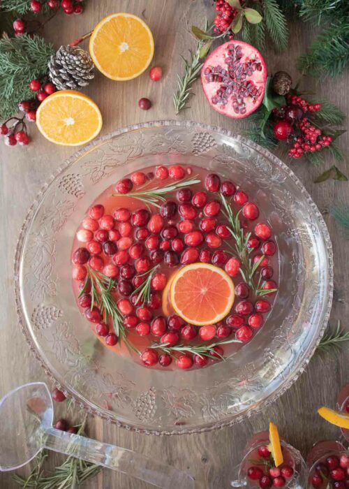 Punch bowl filled with Christmas punch along with garnish of orange slices, cranberries, and rosemary