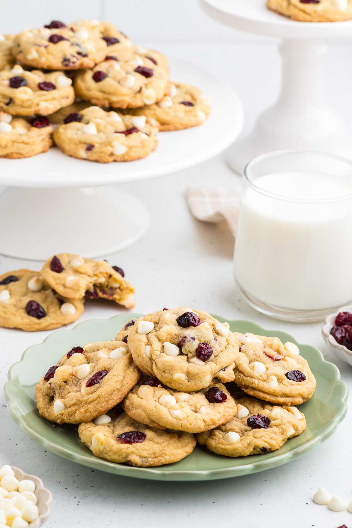 A green plate with Cranberry Orange White Chocolate Chip Cookies and two cookies plus a glass of milk in the background