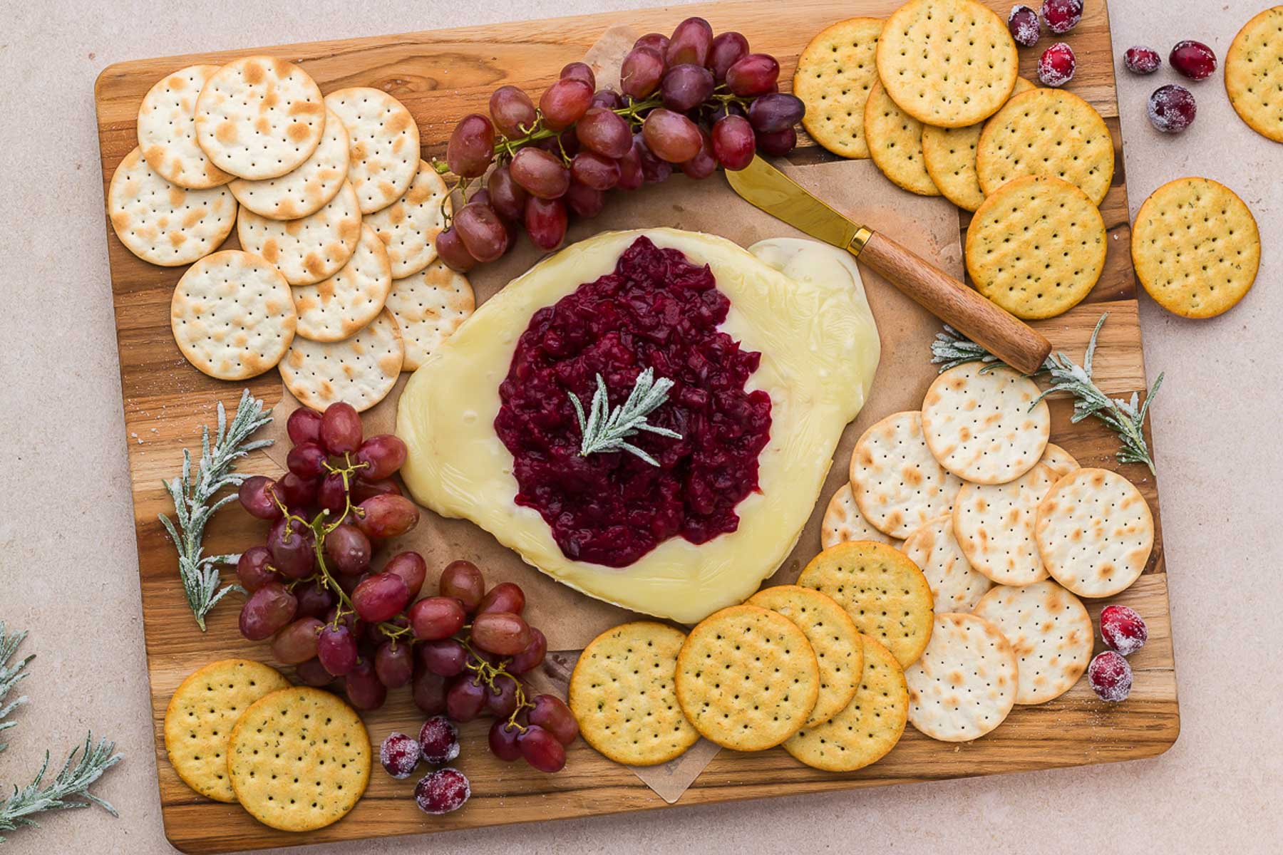A wooden board with warm cranberry baked brie, crackers, and grapes