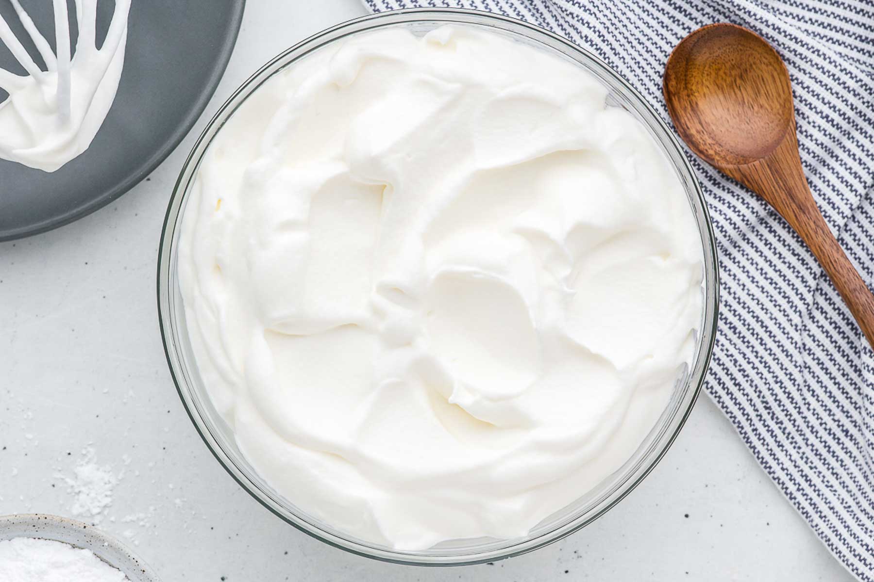 A top view of a bowl of whipped cream with a wooden spoon on a striped napkin.