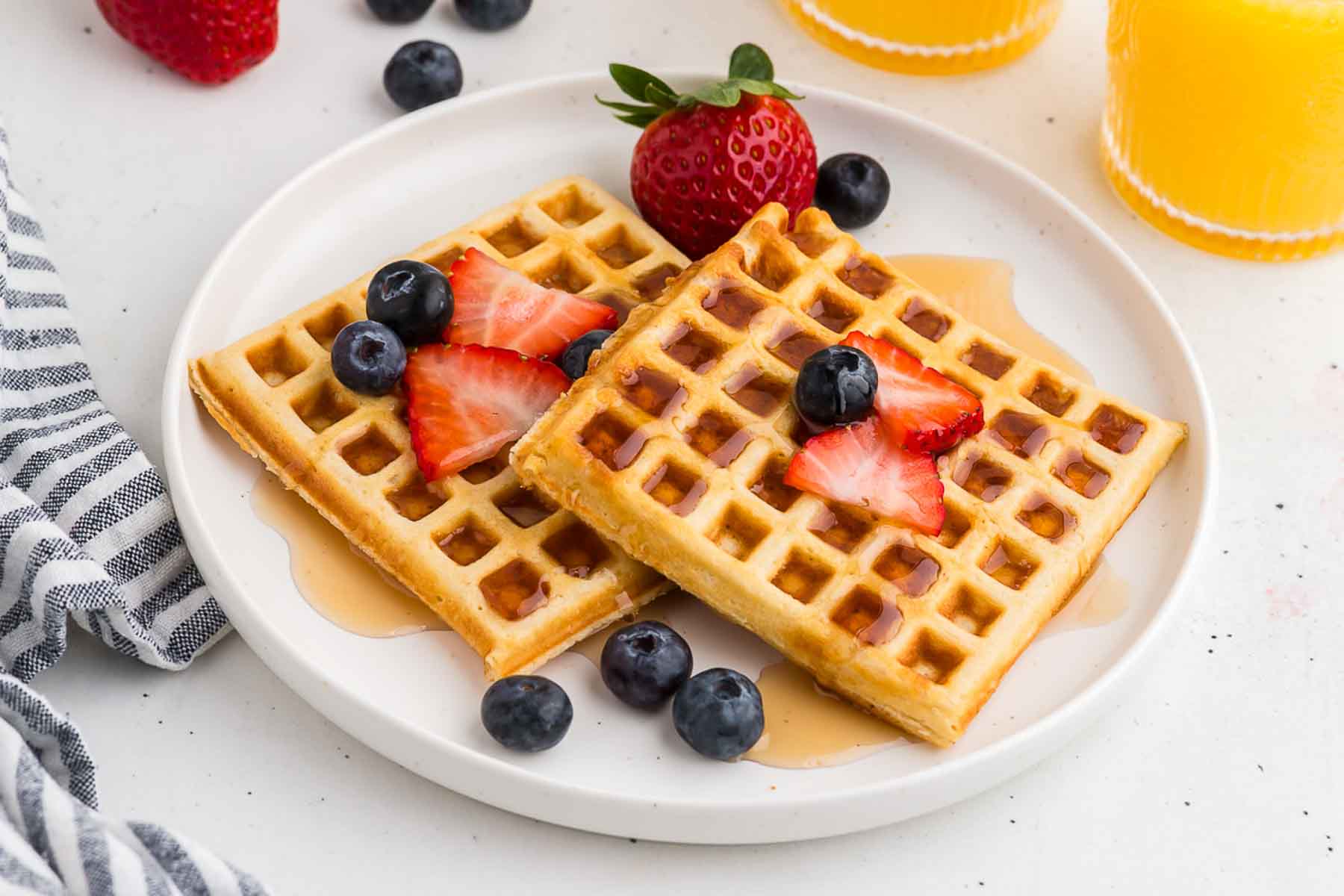 Two golden waffles on a white plate with berries and orange juice in the background