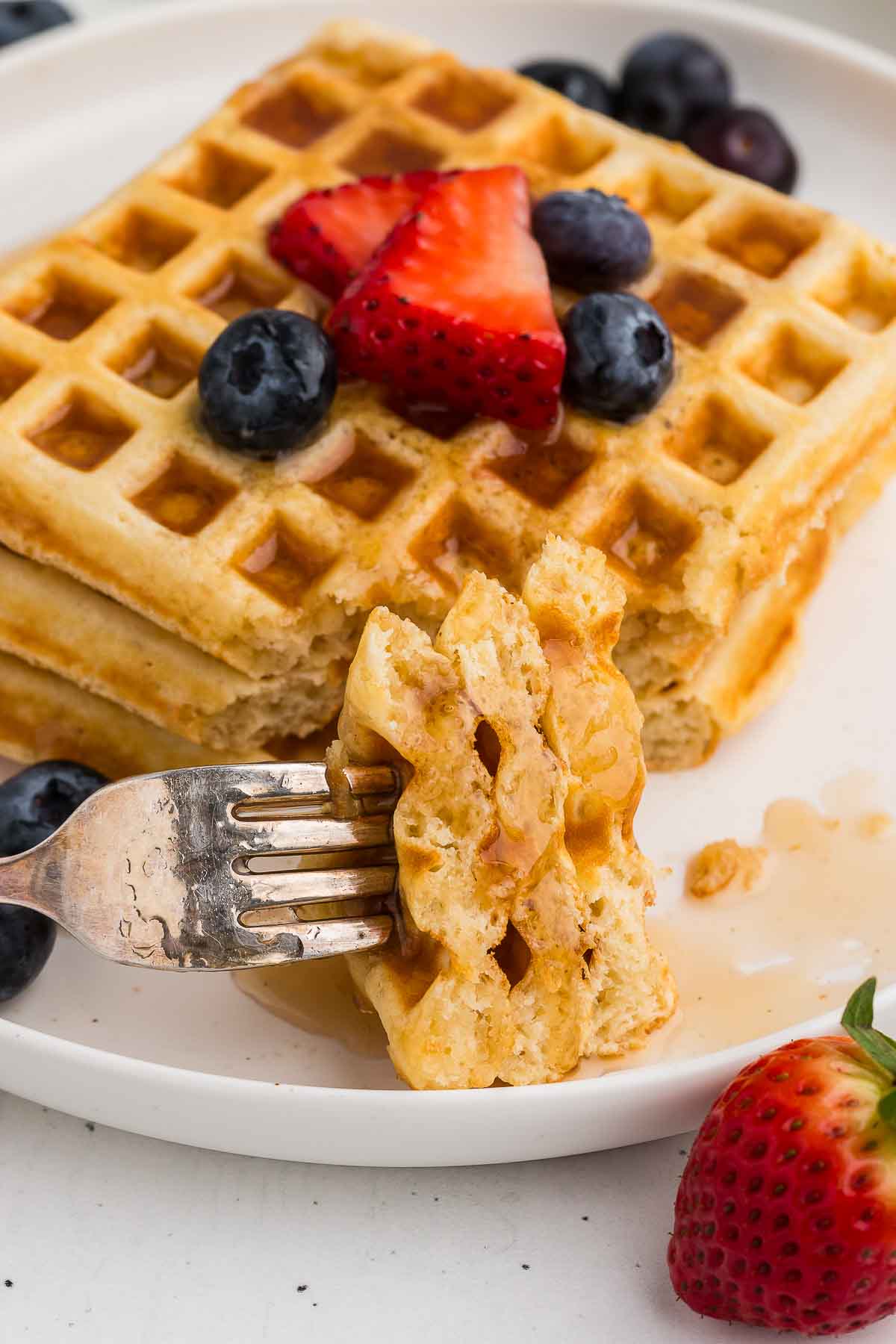 A bite of waffles on a silver fork with a pile of waffles with berries in the background