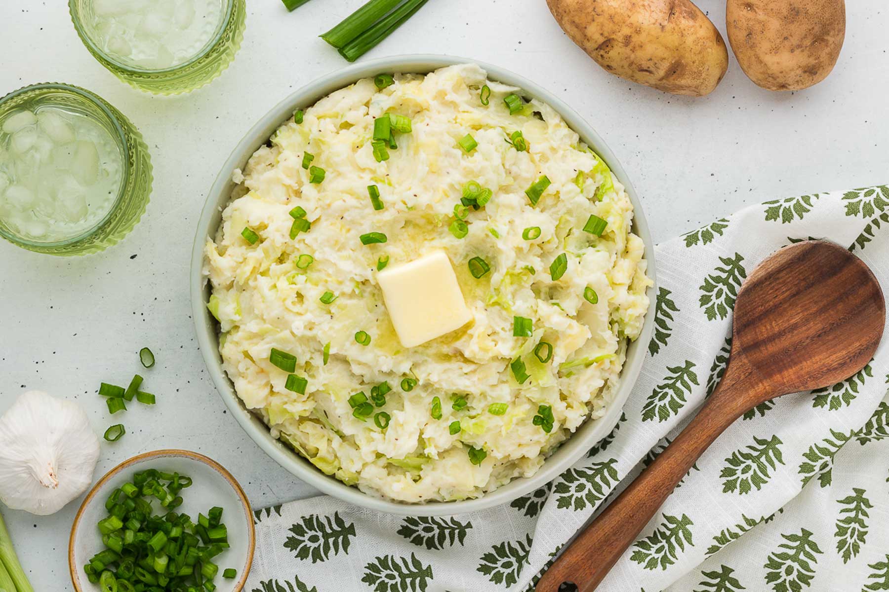 A white bowl full of colcannon with a wooden spoon, green glasses, and potatoes in the background