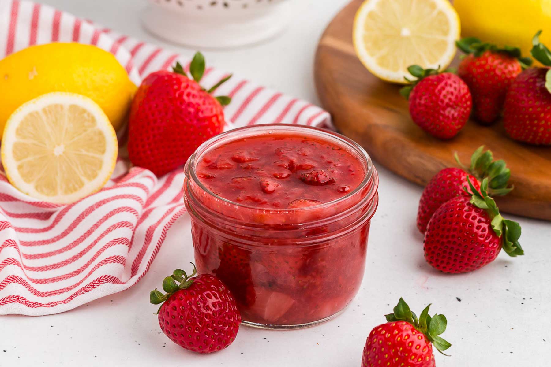 A mason jar with strawberry sauce surrounded by fresh strawberries and lemons on a red and white striped towel