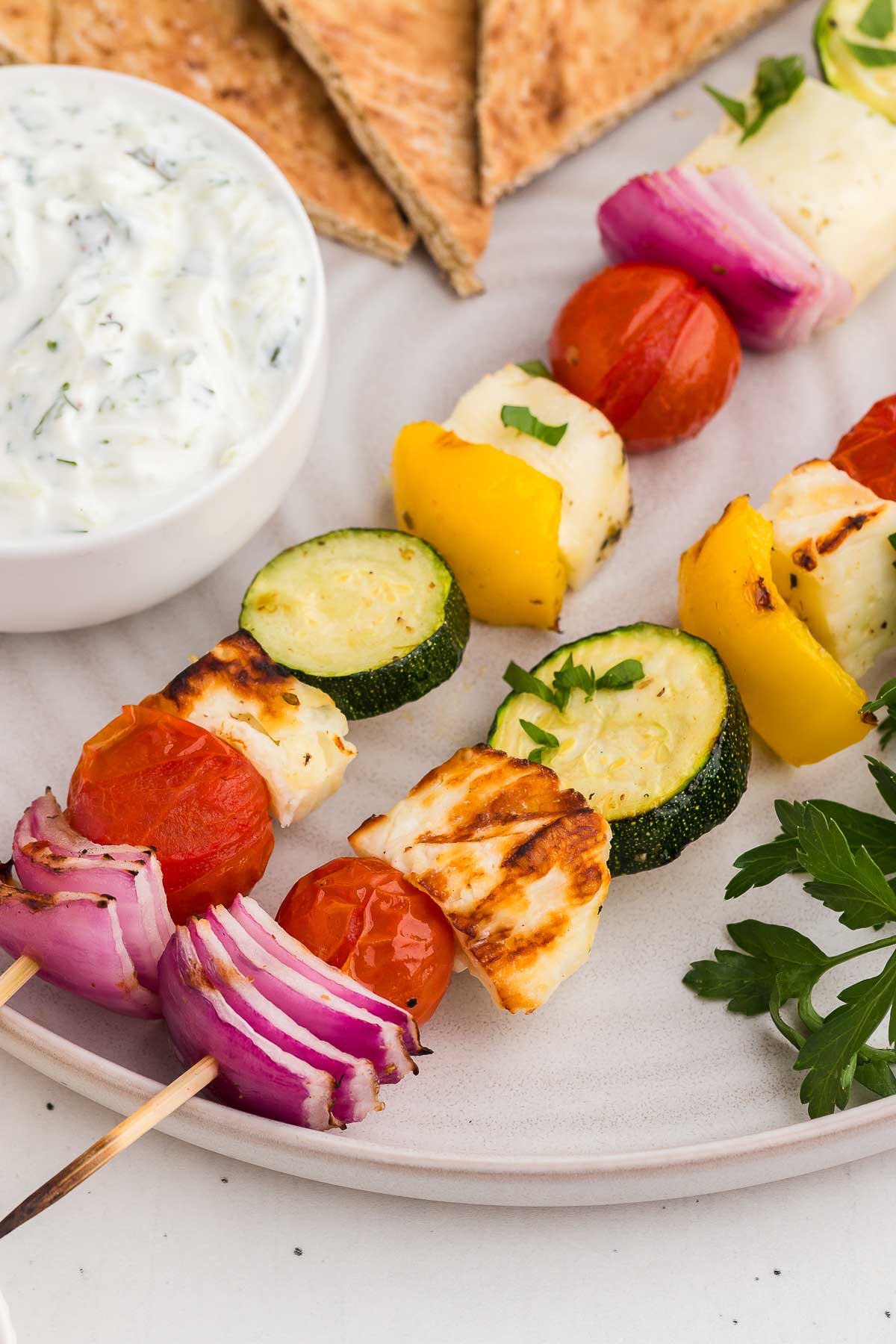 Two grilled halloumi and veg combos on a white plate with herbs, tzatziki sauce, and pita
