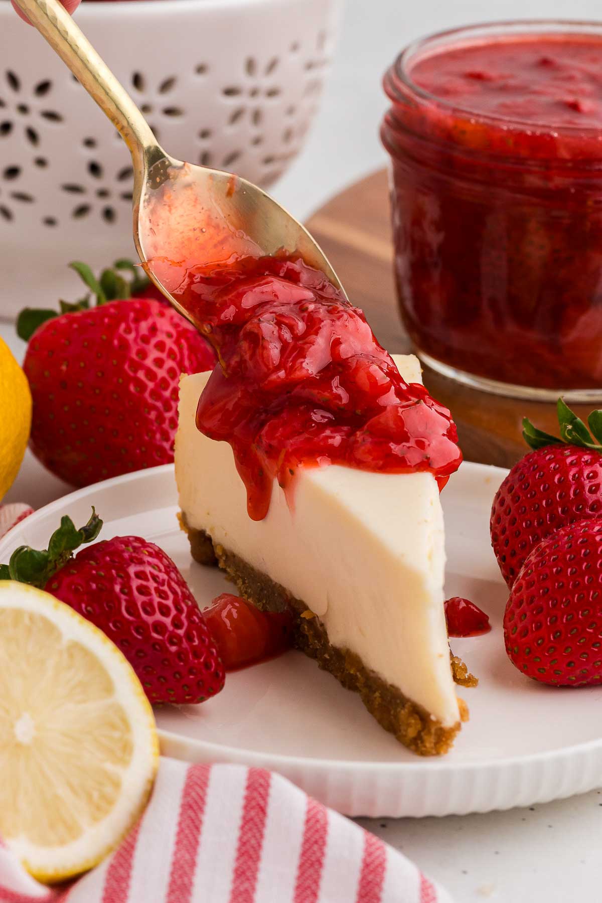 a spoon scoops strawberry sauce over cheesecake on a white plate.