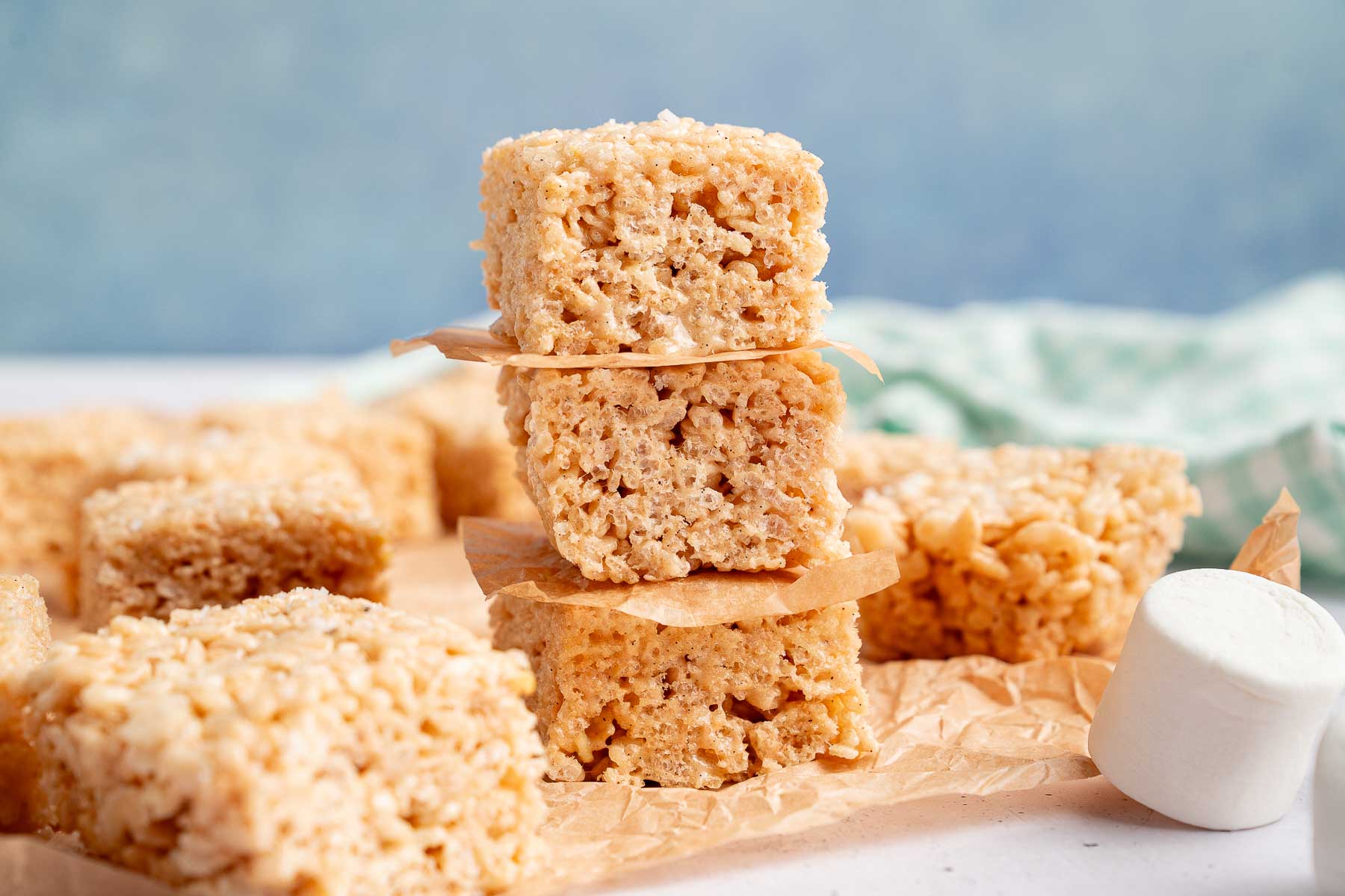 A stack of three Rice Krispie treats with a marshmallow and more treats in the foreground